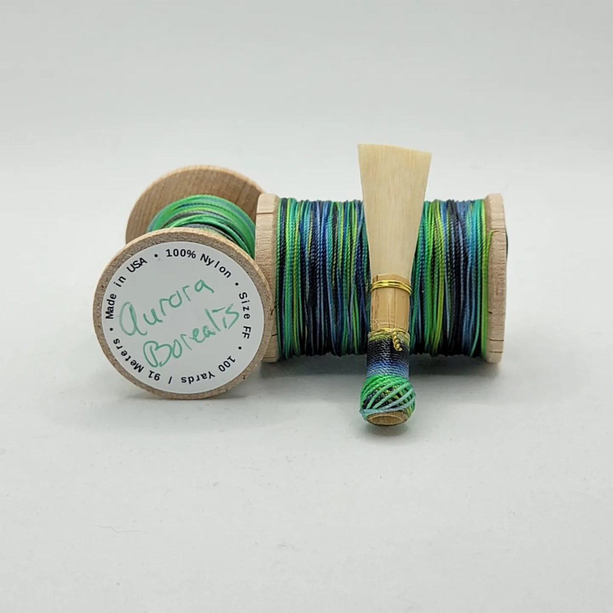 A handcrafted, finished bassoon reed with green, blue, and black thread, displayed on a white background.