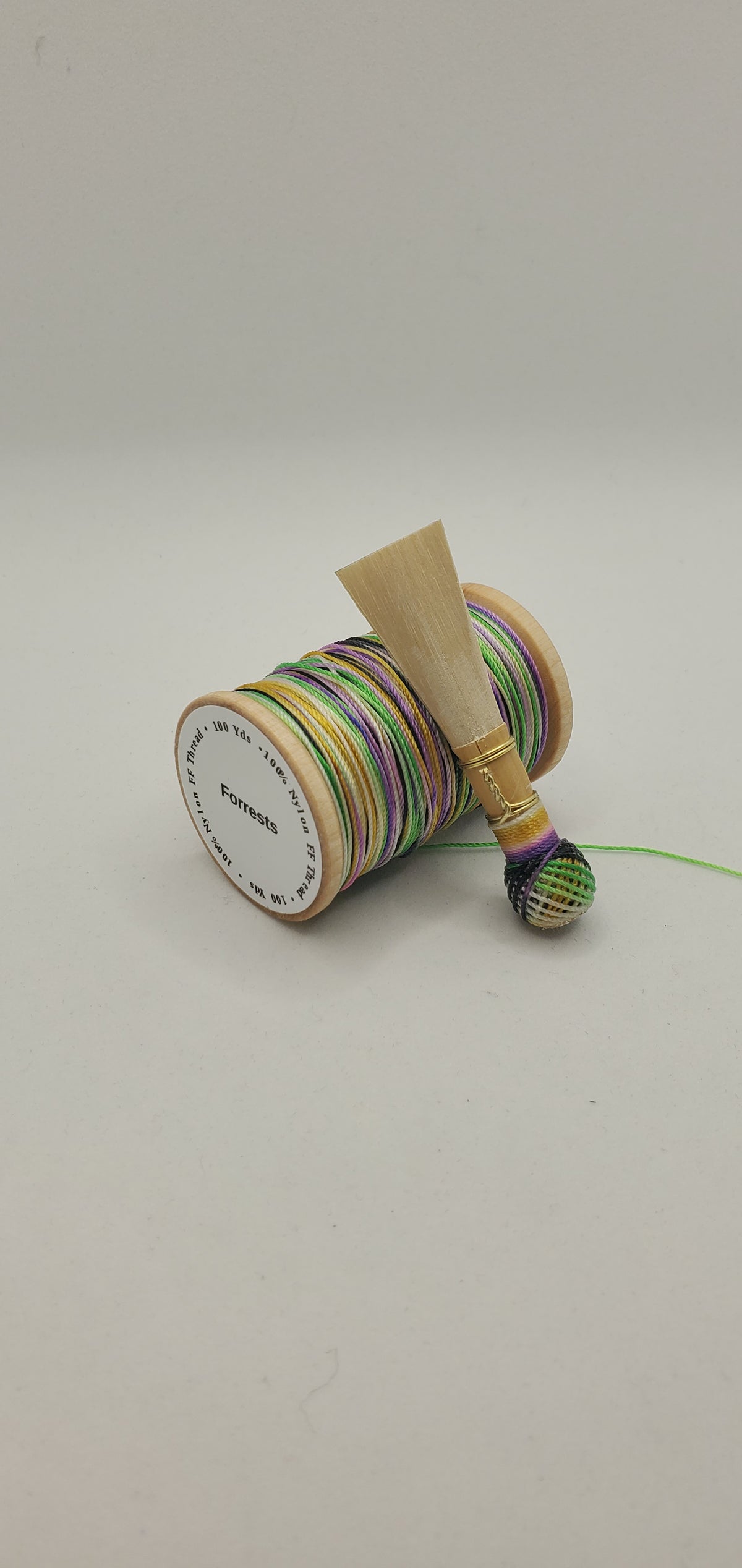 A handcrafted, finished bassoon reed with green, yellow, and purple thread, displayed on a white background.