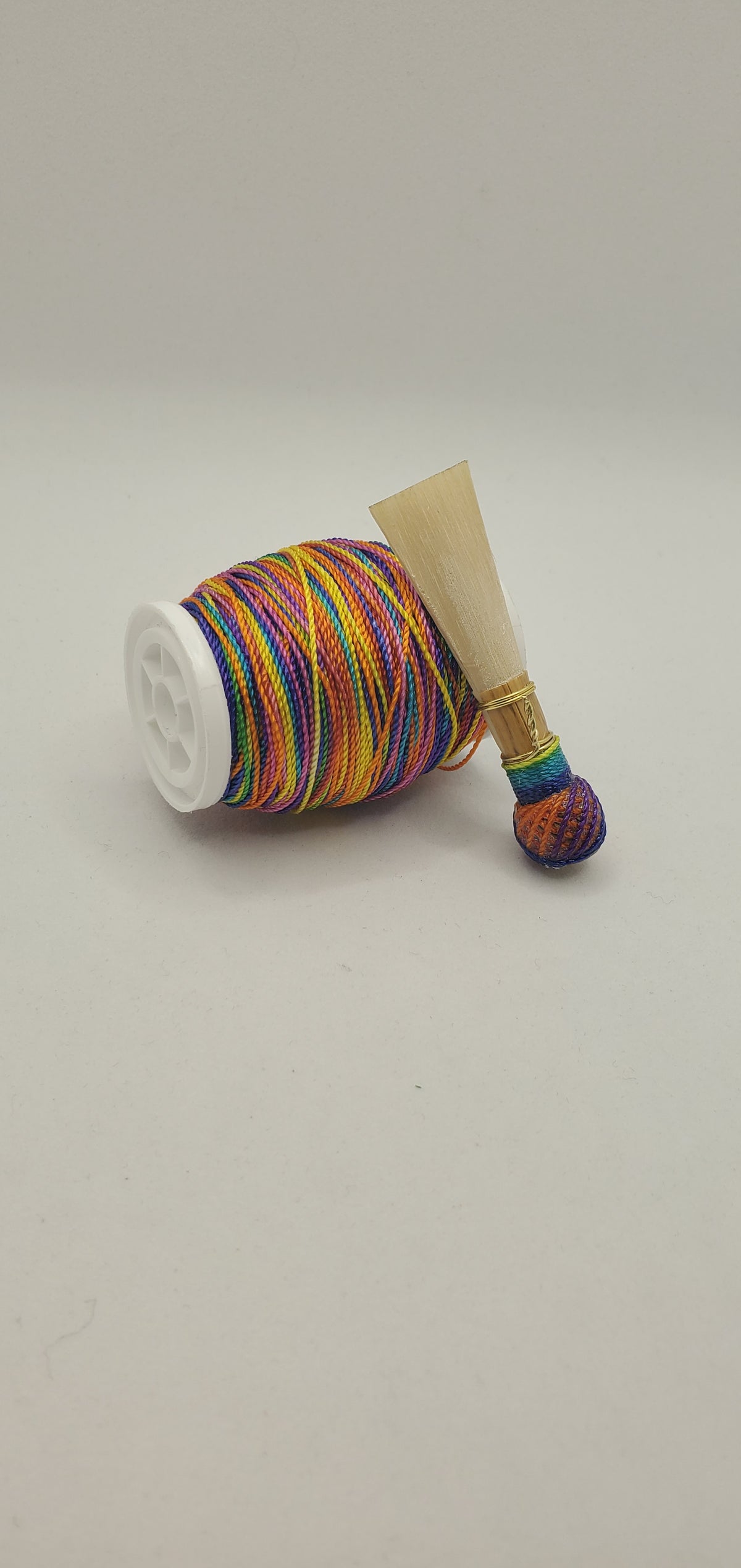A handcrafted, finished bassoon reed with rainbow multicolored thread, displayed on a white background.