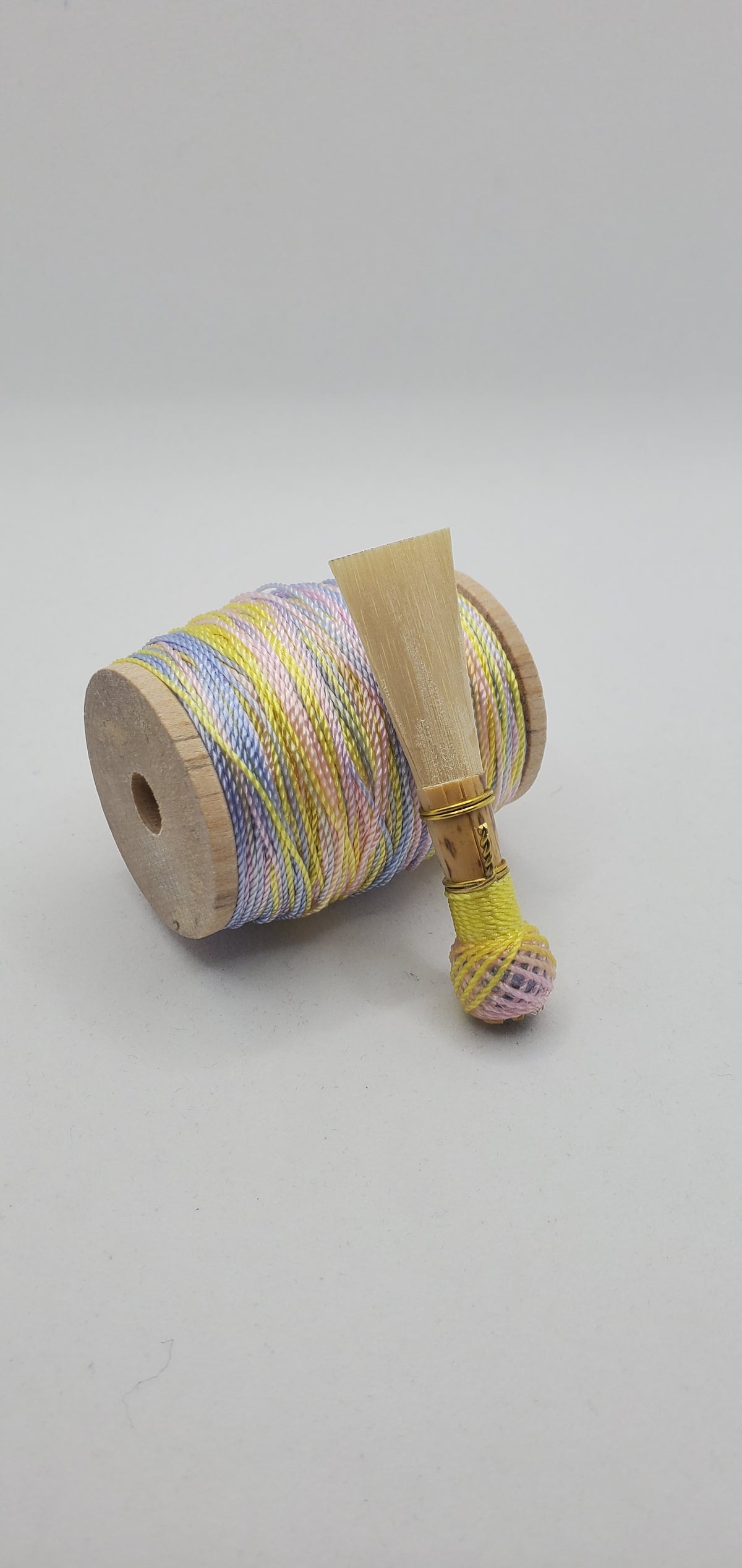 A handcrafted, finished bassoon reed with pastel pink, blue, and yellow thread, displayed on a white background.